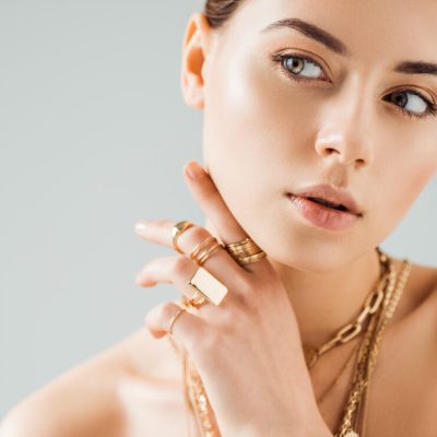 young nude woman in golden rings and necklaces looking away isolated on grey